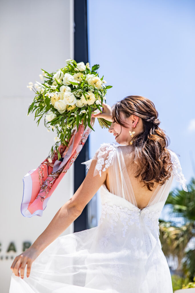 【名古屋市結婚式場ビアンカーラテラス】の開放的な空間にて、青空を背に笑顔で振り返る美しいドレス姿の花嫁様。名古屋市にある市の中心部で叶える、光と風を感じる屋上テラスでのロケーションフォトは、名古屋市内でも開放感あふれる結婚式を求めるお二人の幸せな瞬間を詳細に描写しています。