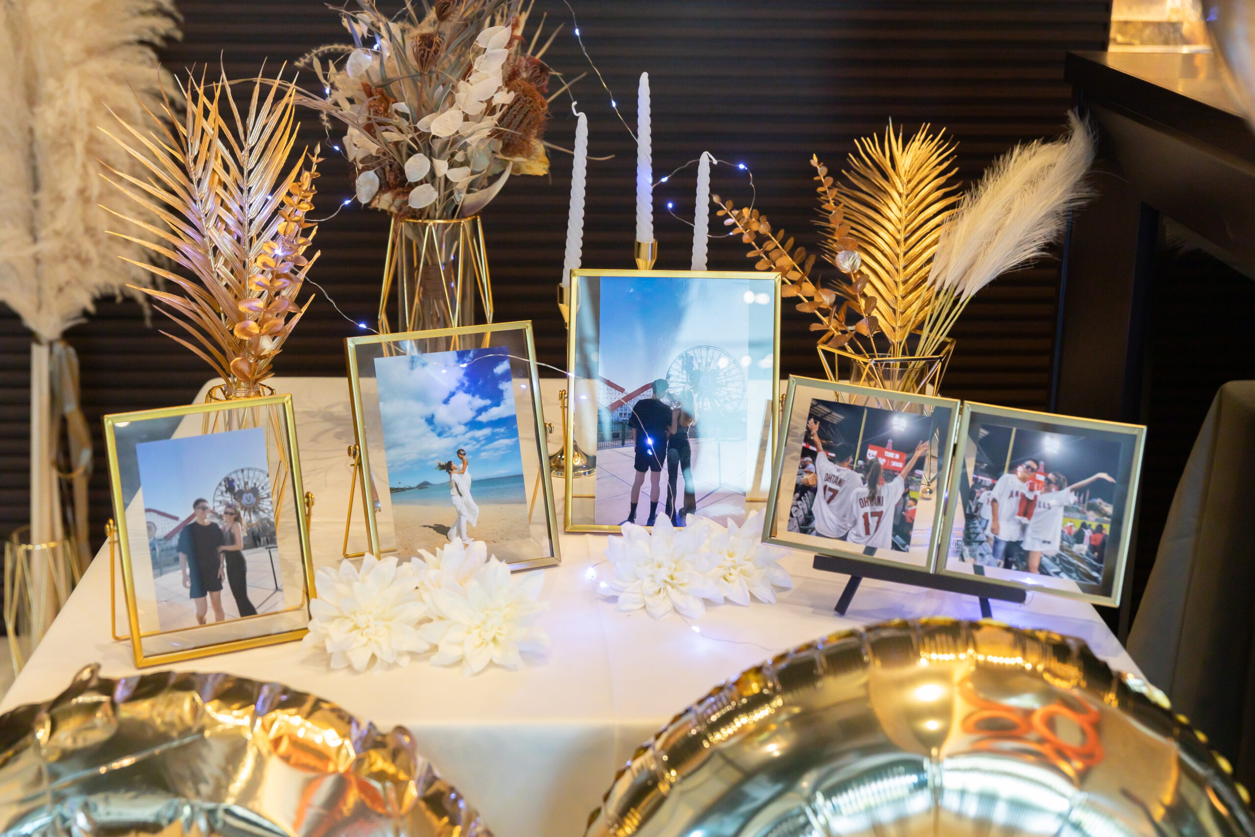 【名古屋市結婚式場ビアンカーラテラス】ゴールドのフレームに収められた、ビーチや遊園地での思い出深い写真たちが並ぶウェルカムスペース。名古屋市にある市の中心部で叶える、お二人らしさを表現したこだわりのおもてなし空間は、名古屋市内での披露宴を待つゲストの皆様を楽しませる、上質で温かな情景を詳細に描写しています。