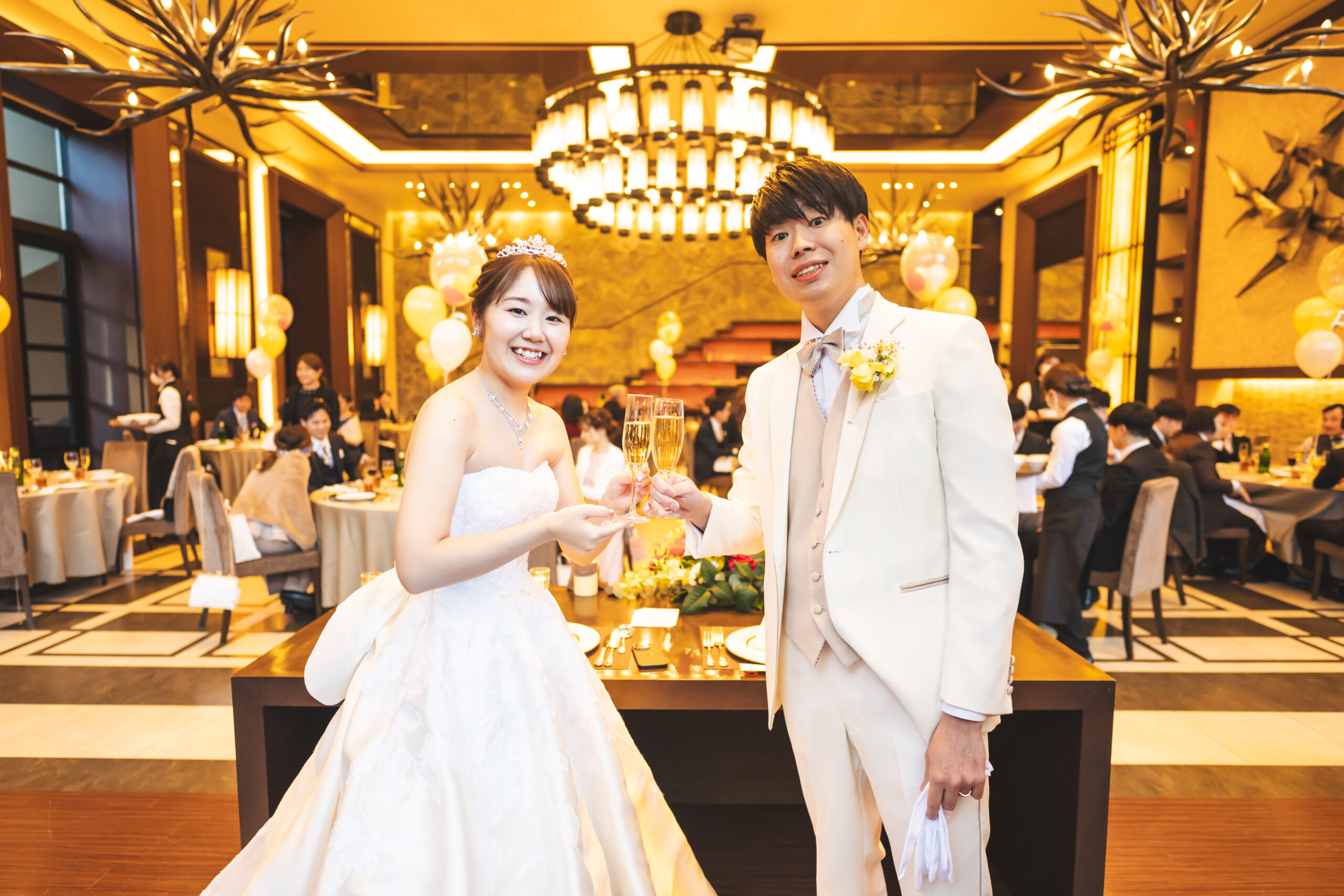 【名古屋市結婚式場ビアンカーラテラス】豪華なシャンデリアが輝くモダンな披露宴会場で、笑顔で乾杯を交わす幸せいっぱいな新郎新婦様。名古屋市にある市の中心部という好立地を活かし、大切なゲストを招待して華やかでアットホームなパーティーを楽しむ、一生の思い出に残るウェディングシーンを象徴する一枚です。