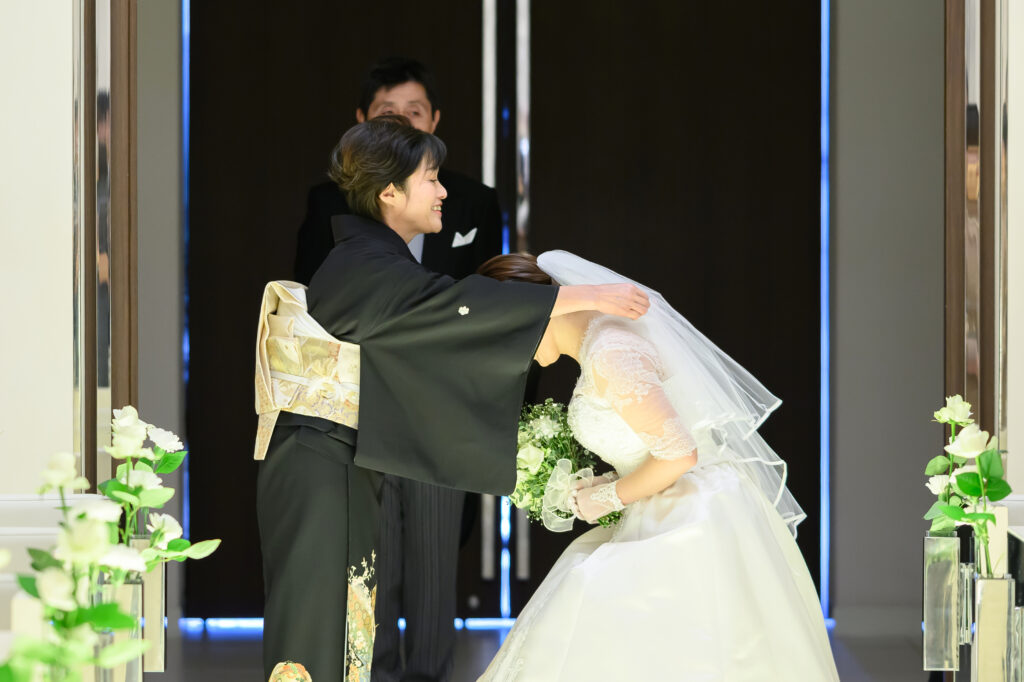 【名古屋市結婚式場ビアンカーラテラス】挙式の直前、お母様が優しく新婦様のベールを下ろす感動的なベールダウンの瞬間。名古屋市にある市の中心部で叶える、親子の絆を再確認するこの神聖な儀式は、名古屋市内での挙式を控えた多くの花嫁様が憧れる、一生の思い出に残る美しい情景を詳細に描写しています。