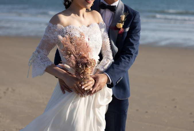 【名古屋市結婚式場ビアンカーラテラス】穏やかな波が寄せるビーチをバックに、繊細なレースの袖が美しいドレスを纏った花嫁様を新郎様が後ろから抱きしめるロマンティックなショット。名古屋市にある市の中心部から離れ、開放感あふれる海辺で撮影するロケーションフォトは、名古屋市内での結婚式に彩りを添える一生の思い出となる一枚です。