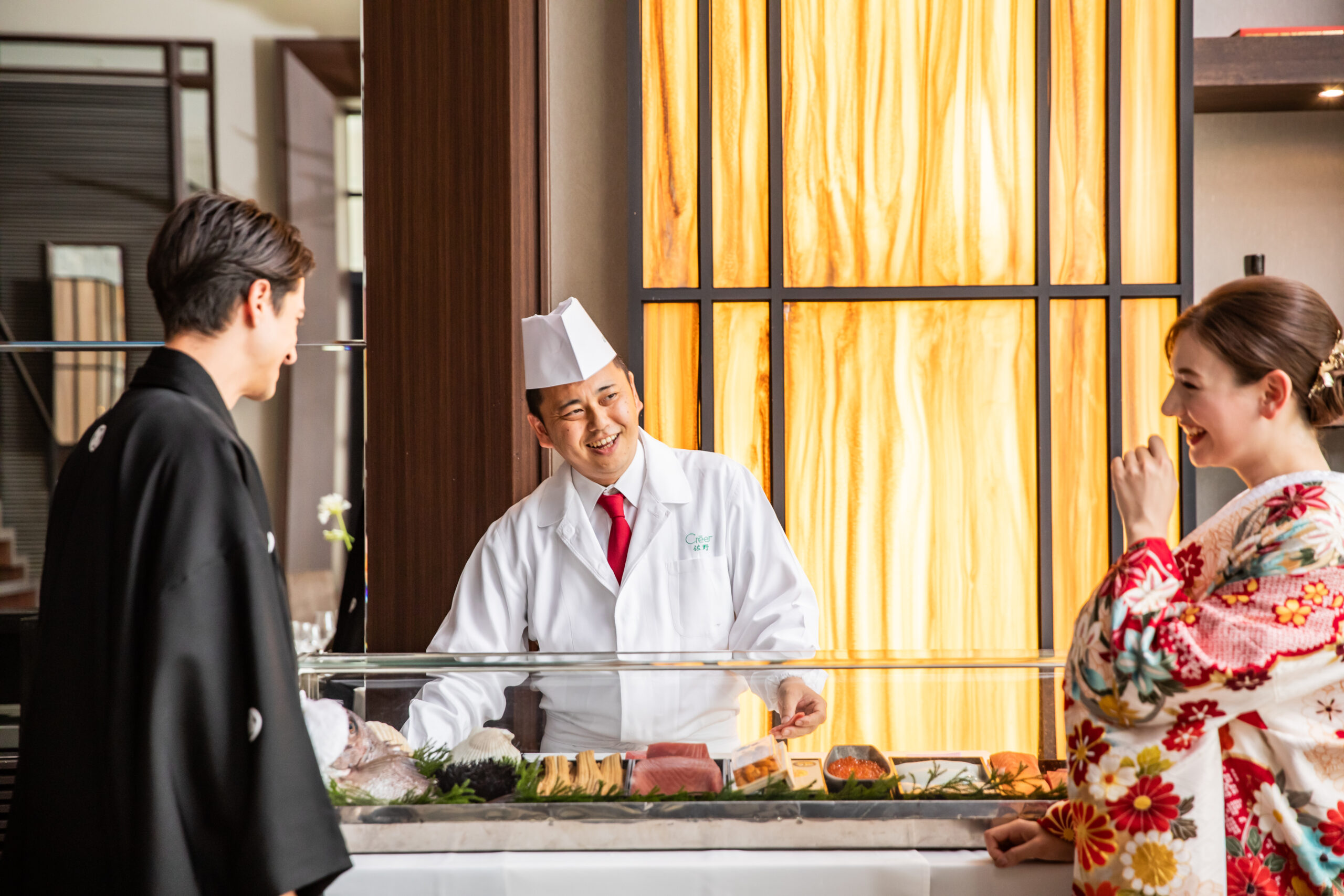 【名古屋市結婚式場ビアンカーラテラス】披露宴会場内に設けられた特設カウンターで、職人が握る本格的な寿司ビュッフェを楽しむ和装姿の新郎新婦様。名古屋市にある市の中心部で叶える、美食にこだわったおもてなし演出は、名古屋市内外から訪れるゲストの皆様に大変喜ばれる特別なプログラムであることを詳細に描写しています。