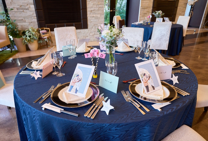 【名古屋市結婚式場ビアンカーラテラス】の披露宴会場で、ネイビーのクロスにパステルカラーの席札が映えるテーブルコーディネート。名古屋市にある市の中心部で叶える披露宴では、お二人の写真を活用したペーパーアイテムでゲストを詳細に描写し、感謝を伝える温かなおもてなしの空間を演出しています。