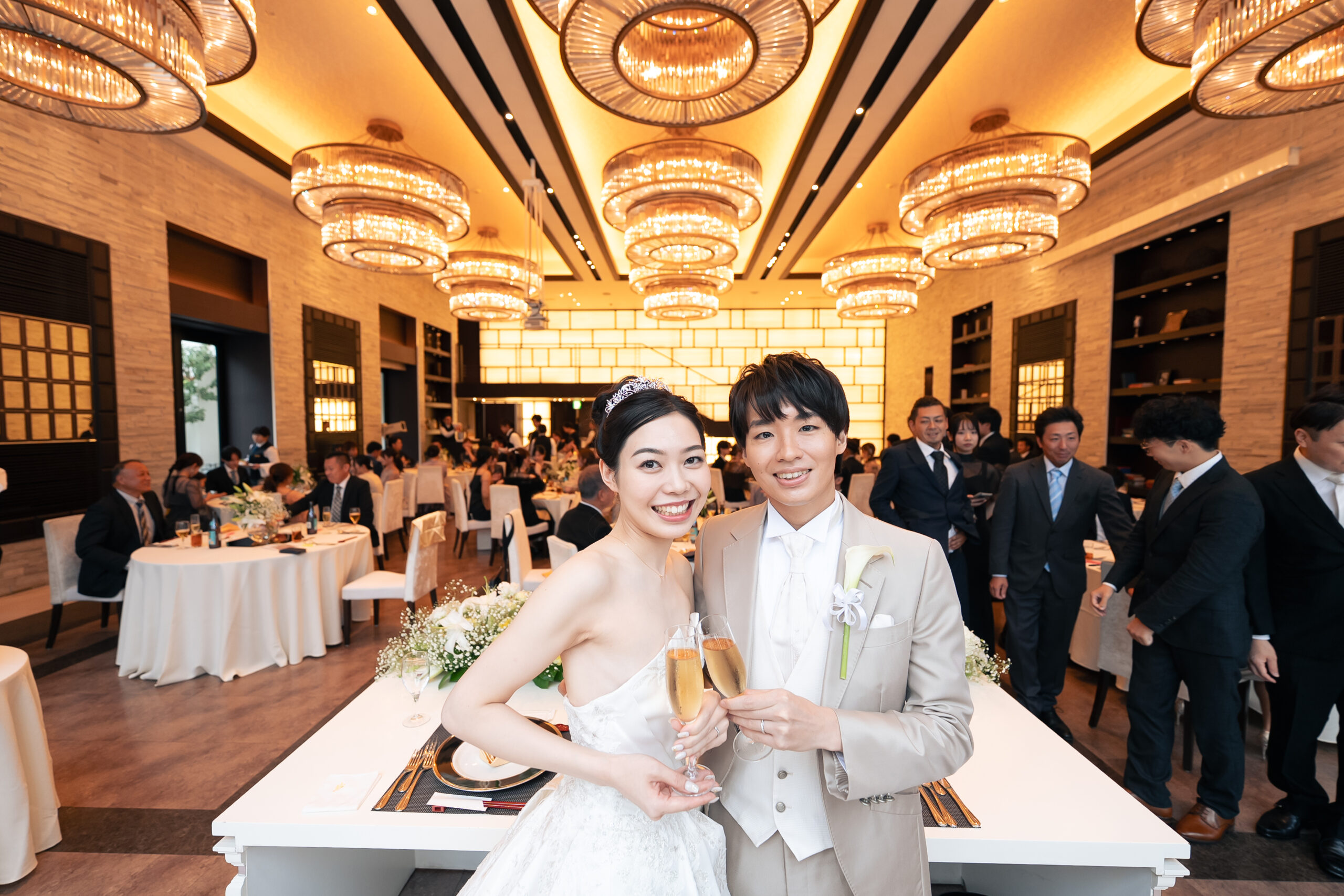 名古屋市の結婚式場ビアンカーラヒルサイドテラス。豪華なシャンデリアが輝く披露宴会場で乾杯する新郎新婦。ゲストの笑顔に囲まれた華やかなパーティー。