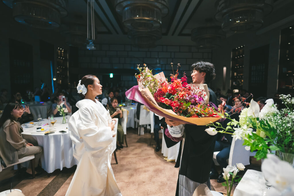 名古屋市の結婚式場ビアンカーラヒルサイドテラス。白無垢姿の新婦へ、新郎がサプライズで巨大な赤のブーケを贈る、和装での感動的な披露宴演出シーン。