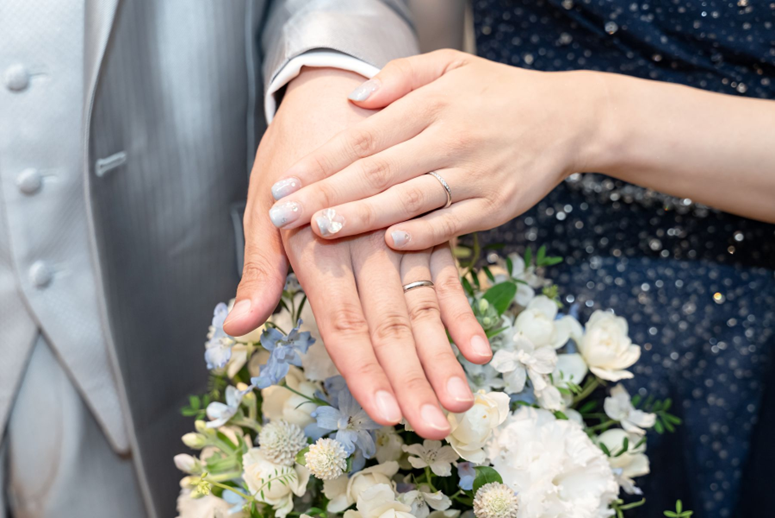 名古屋市結婚式場ビアンカーラヒルサイドテラス。ブルーとホワイトのブーケを背景に、重ね合わせた新郎新婦の手。きらめく結婚指輪と花嫁の繊細なウェディングネイルが美しい、幸せの誓いを象徴するハンドショット。