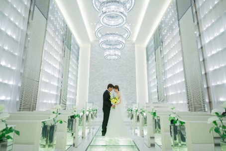 名古屋市結婚式場ビアンカーラヒルサイドテラス。クリスタルが輝くガラスのバージンロードに立つ新郎新婦の後ろ姿。5連のリング型シャンデリアと700個のキャンドルが灯る、ビアンカーラ・ヒルサイドテラスの幻想的なチャペル。