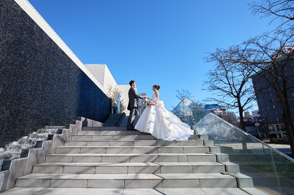 澄み渡る青空の下、名古屋市結婚式場ビアンカーラ・ヒルサイドテラスの象徴的な大階段で手を取り合う新郎新婦。水が流れる壁面とモダンな外観が背景に広がる、開放的なロケーションフォト。