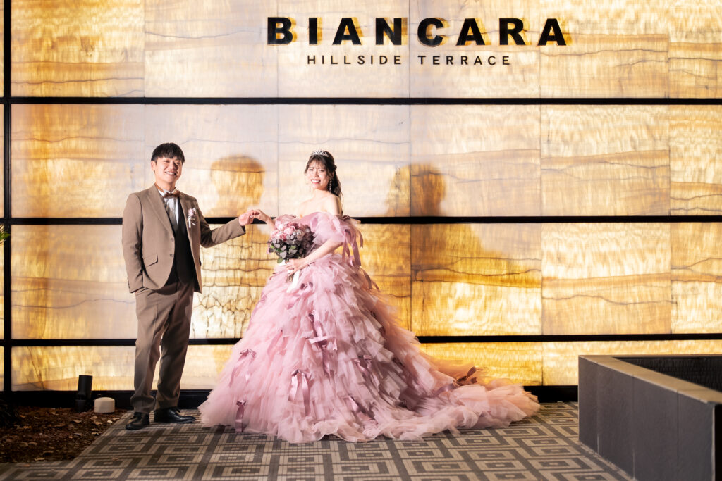 名古屋市の結婚式場ビアンカーラヒルサイドテラスのロゴ前で、華やかなピンクのカラードレス姿の新婦と新郎が手を取り合い笑顔を見せるウェディングフォト