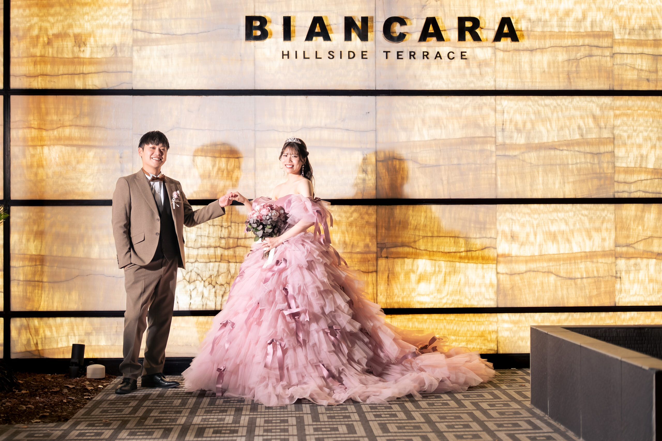 名古屋市の結婚式場ビアンカーラヒルサイドテラスのロゴ前で、華やかなピンクのカラードレス姿の新婦と新郎が手を取り合い笑顔を見せるウェディングフォト