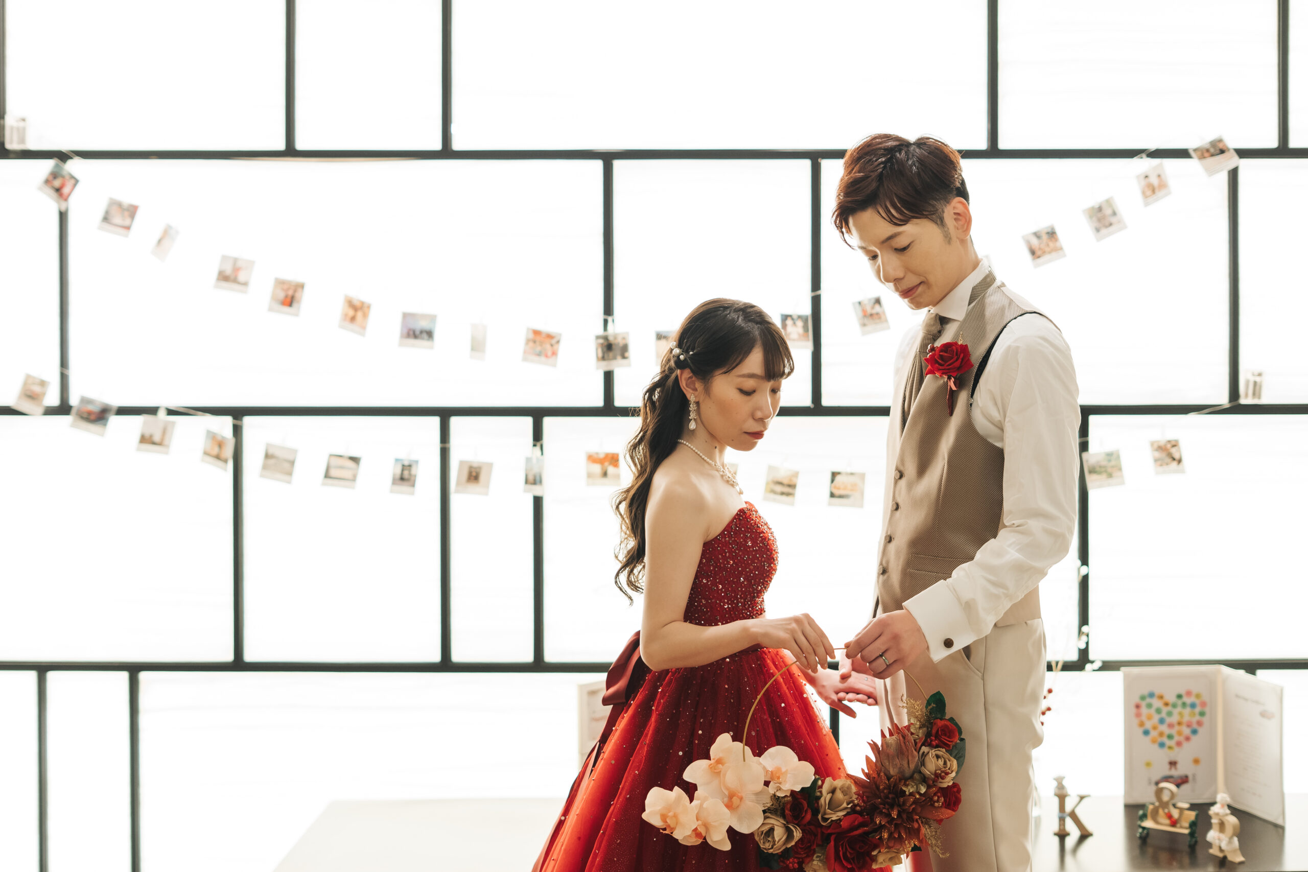 名古屋市の結婚式場ビアンカーラヒルサイドテラスの披露宴会場で、赤いカラードレスの新婦と新郎が、背景のフォトガーランドの前でリースブーケを手にする様子
