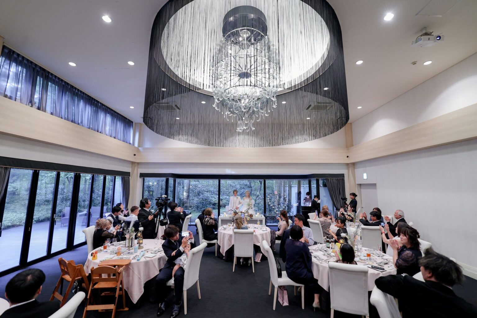 名古屋市の結婚式場ビアンカーラヒルサイドテラスの開放感溢れる披露宴会場で、大きなシャンデリアの下、ゲストの祝福に包まれるメインテーブルの新郎新婦