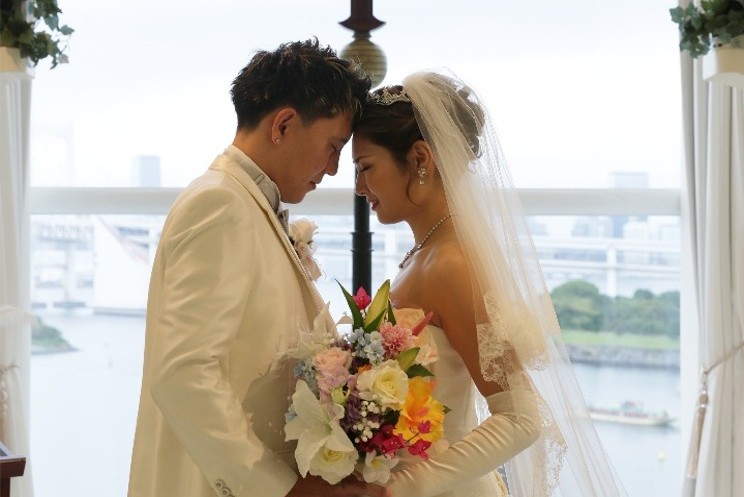 名古屋市の結婚式場ビアンカーラヒルサイドテラスのチャペル。逆光の中、額を寄せ合う新郎新婦と色鮮やかなブーケ。背景の窓からは十字架と、美しい水辺の景色が広がる。