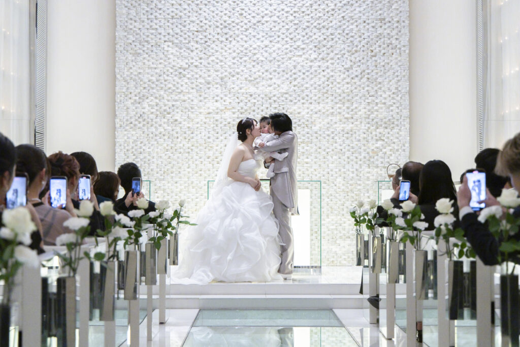 白亜のチャペルにて、お子様を囲んで幸せなキスを交わす新郎新婦様です。ガラスのバージンロードが輝く幻想的な空間で、大切なゲストに見守られながら家族の絆を誓い合う至福のひとときをふんわりと表現しています。