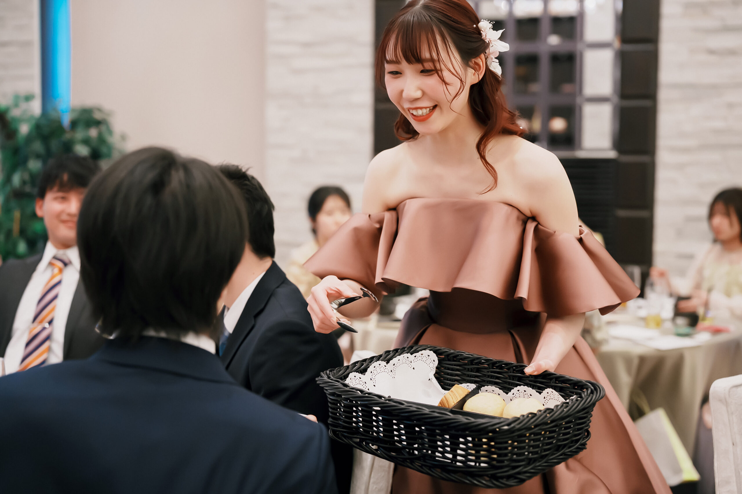 披露宴会場にて、お色直し後の新婦様がゲストの皆様へパンサーブを行うおもてなしシーンです。華やかなドレス姿の新婦様が笑顔でパンを振る舞う姿に、ゲストの皆様との絆が深まるアットホームで至福のひとときがふんわりと表現されています。