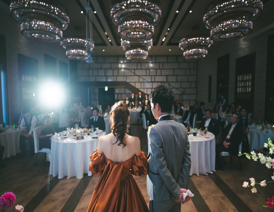 名古屋市の結婚式場ビアンカーラヒルサイドテラスの披露宴。シャンデリアが輝くモダンな会場で、テラコッタカラーのドレスを着た新婦と新郎の後ろ姿。