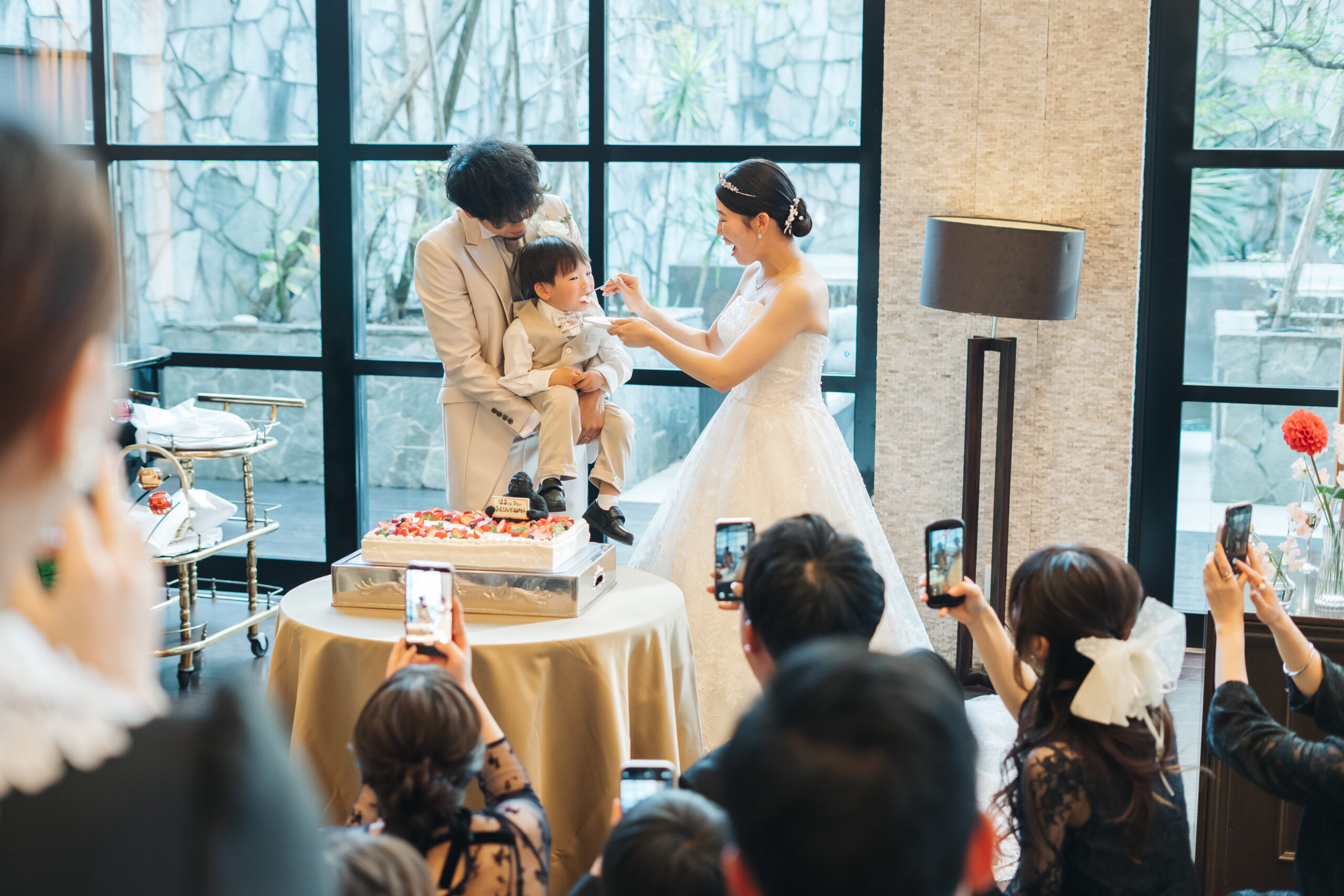 披露宴会場でドレス姿の新婦が新郎に抱かれた子どもへケーキを食べさせるサンクスバイト。
