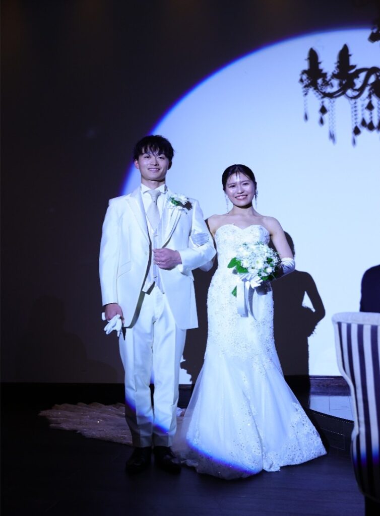 ブライダルフェアの模擬挙式体験。スポットライトに照らされた独立型チャペルで新郎新婦が退場する様子。白いタキシードとマーメイドラインのドレス。