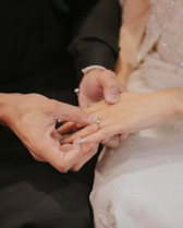 タキシード姿の男性が、ウエディングドレス姿の女性の左手薬指にダイヤモンドの指輪を優しくそっとはめる、結婚式や記念日の指輪交換の手元クローズアップ写真。
