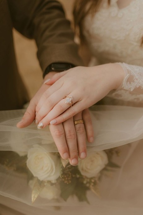 結婚式で白バラのウェディングブーケとベールの上に手を重ねる新郎新婦。ゴールドの結婚指輪と華やかな婚約指輪が輝く花嫁の手元。