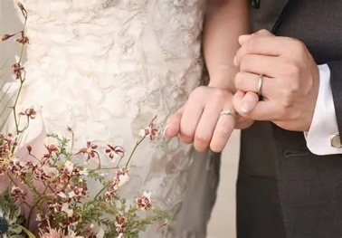 刺繍が施されたウエディングドレスとタキシード姿の新郎新婦が、結婚指輪をはめた手を優しく重ね合わせる、お洒落なウエディングブーケを添えた手元の写真。